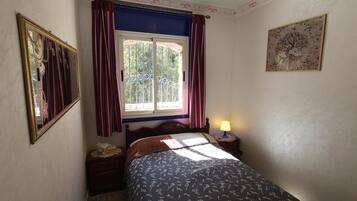 A bedroom with a bed, a nightstand, a lamp, a window with curtains, and a wall-mounted picture.