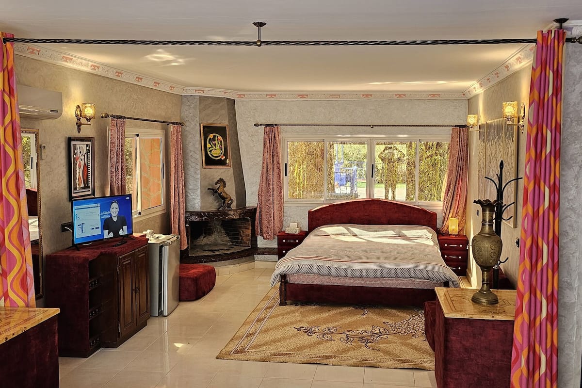 A spacious bedroom with a large bed, a television, a fireplace, and a bathroom with a mirror and sink.