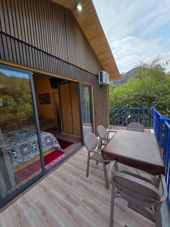 A modern room with a wooden ceiling, a large sliding glass door leading to a balcony, a small bed with a patterned bedspread, and a table with chairs.