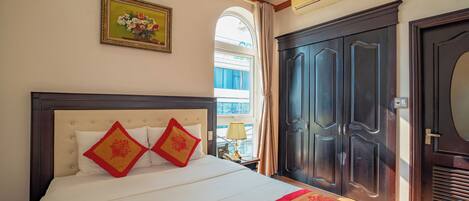 A hotel room with a bed, a large dark wooden wardrobe, a window with curtains, and a framed floral artwork on the wall.