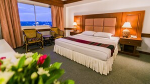 A hotel room with a large bed, two bedside tables with lamps, a chair, a window with curtains, and a view of the cityscape. - Raya Grand Hotel (Nakhon Ratchasima)