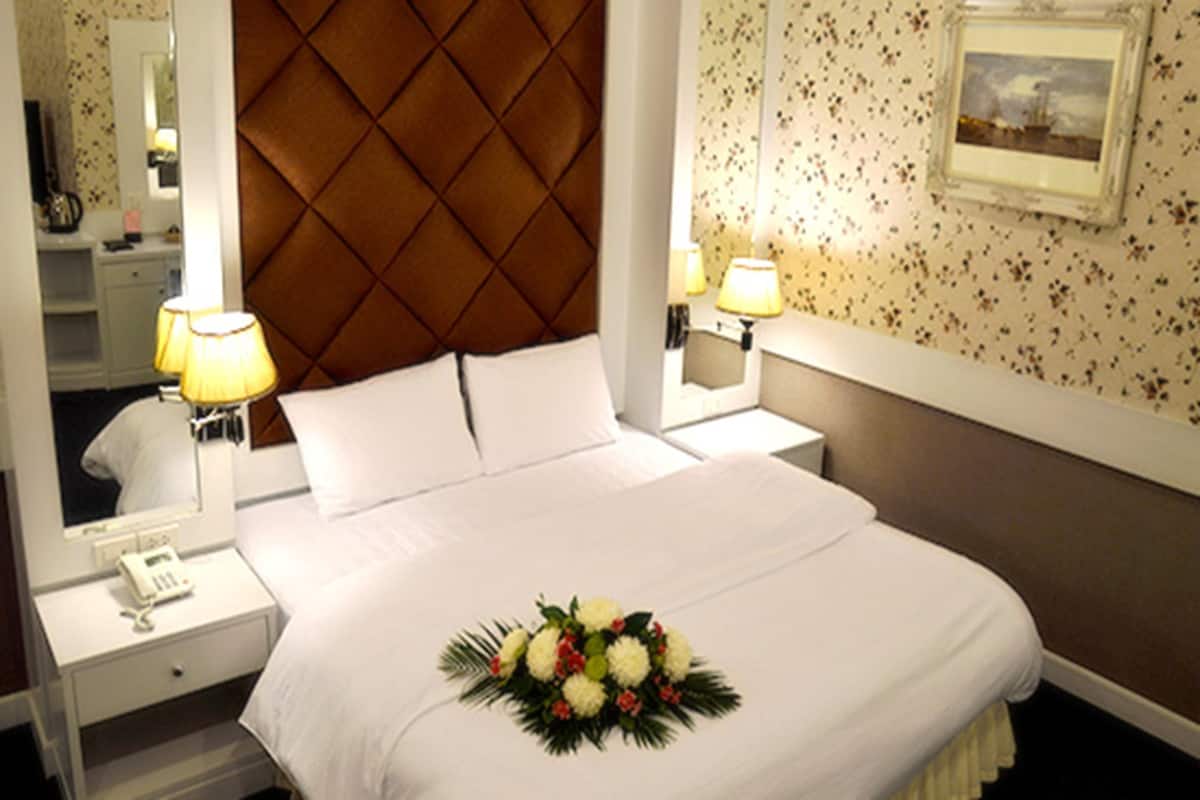 A hotel room with a large bed, a headboard, bedside lamps, a nightstand, a mirror, and a floral arrangement on the bed.