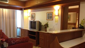 A living room with a television, wooden cabinets, and a large window with curtains. - Raya Grand Hotel (Nakhon Ratchasima)