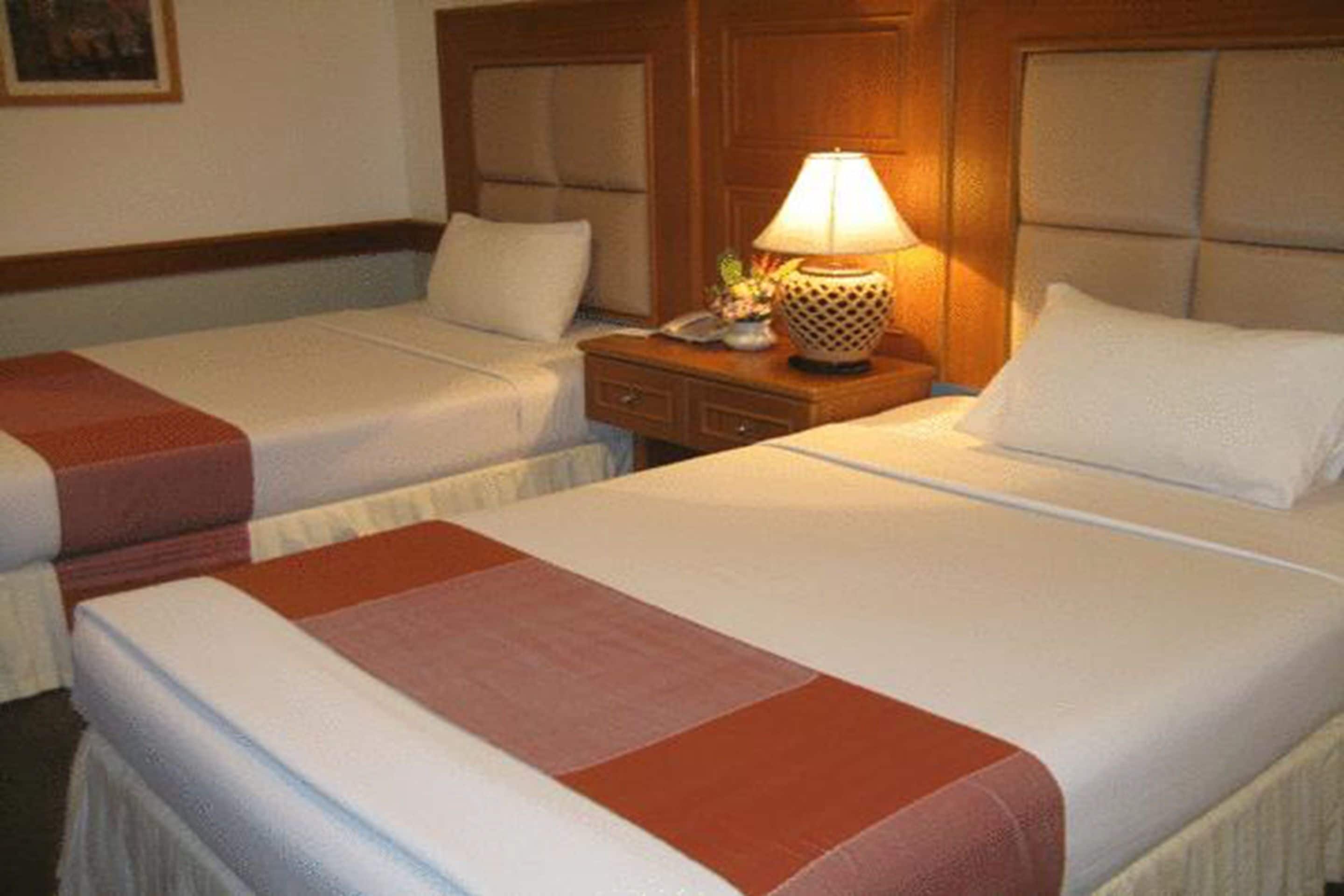 A hotel room with two beds, each with a white bedspread and a red blanket. There is a wooden headboard, a nightstand with a lamp, and a small table with a phone and a decorative item.