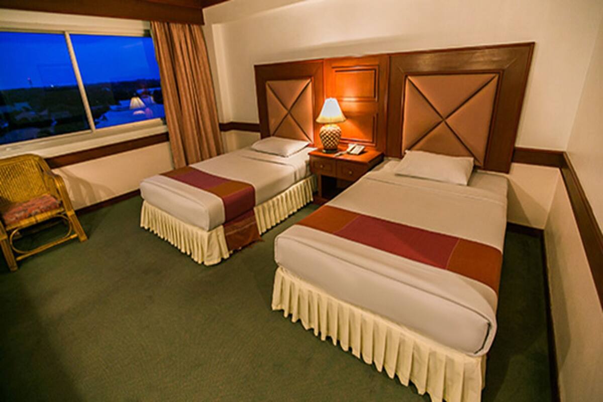 A hotel room with two beds, a nightstand with a lamp, a chair, and a view of the outside.