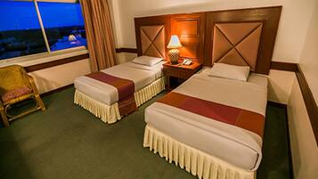 A hotel room with two beds, a nightstand with a lamp, a chair, and a view of the outside.