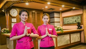 Two receptionists in pink uniforms standing in a lobby with wooden paneling, a clock, and a framed artwork on the wall. - Raya Grand Hotel (Nakhon Ratchasima)