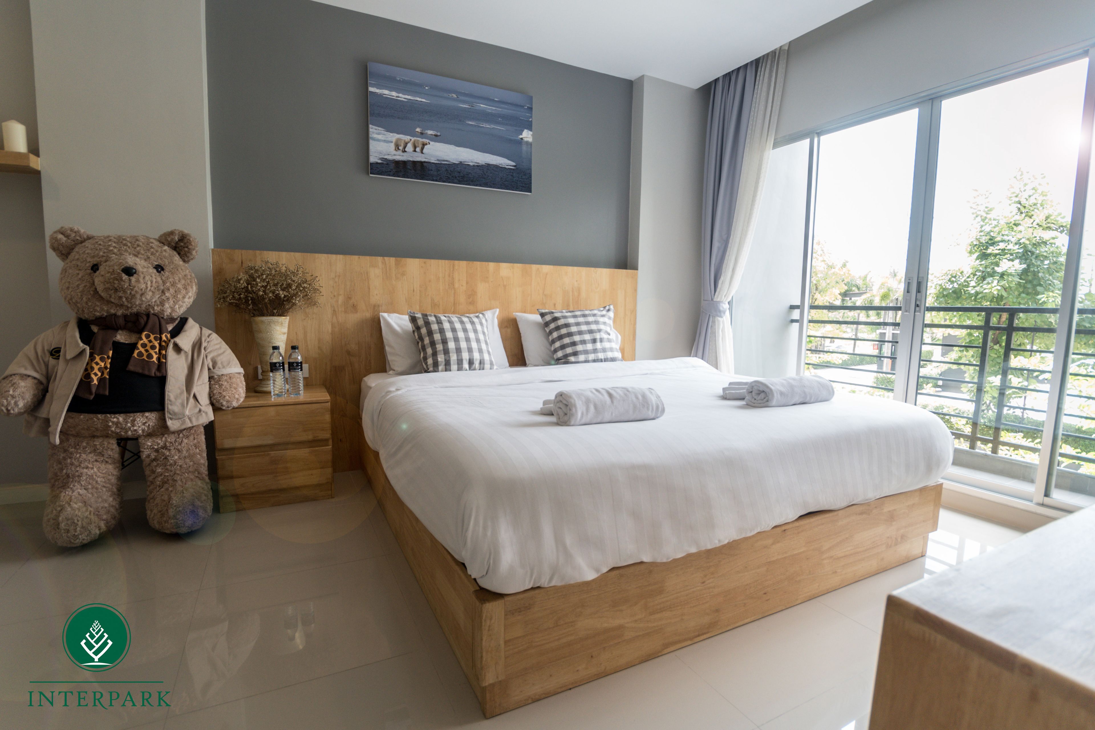 A modern bedroom with a large bed, a wooden headboard, a nightstand with a teddy bear, a potted plant, and a painting of a snowy landscape.