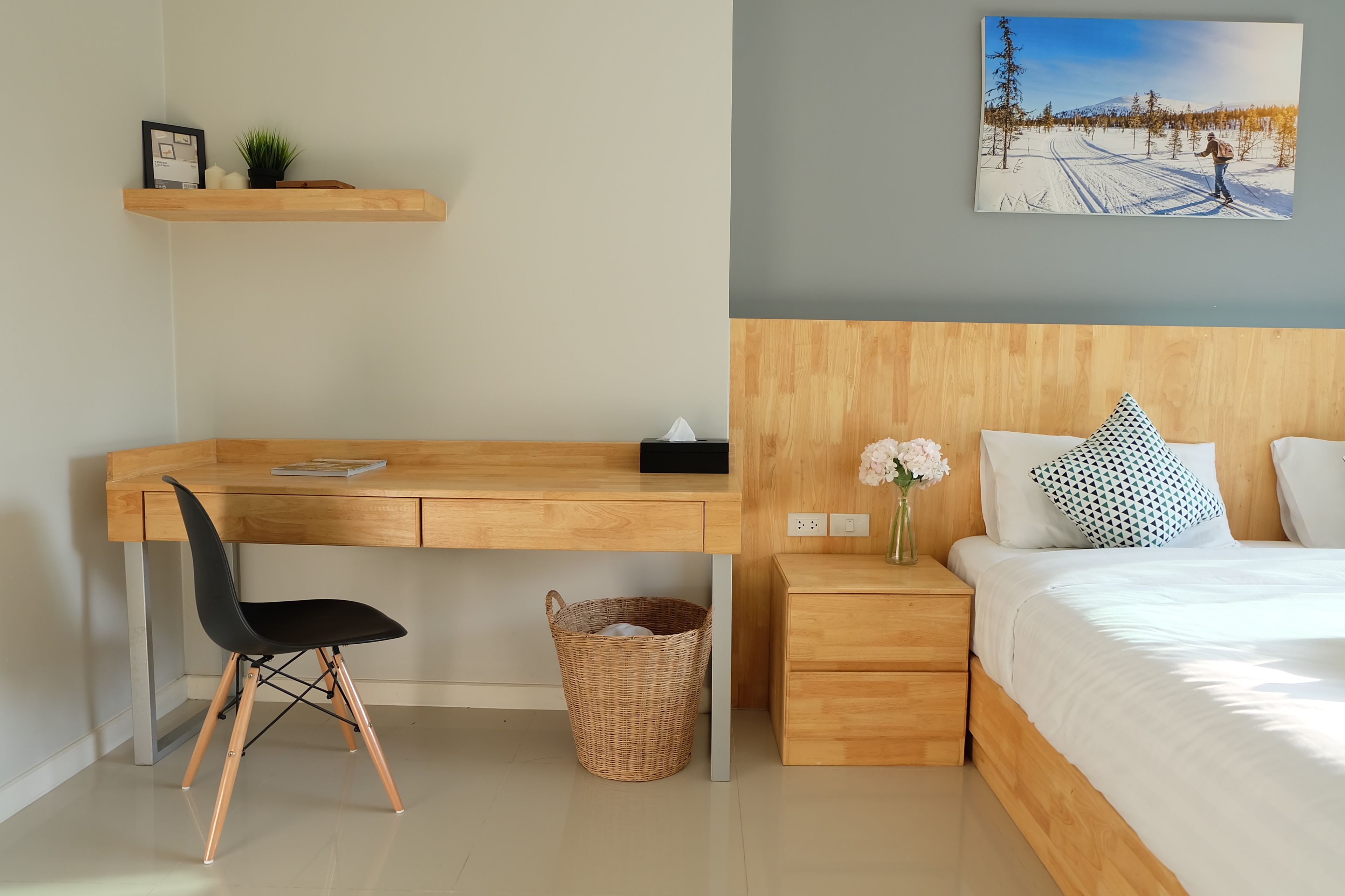 Un dormitorio moderno con un escritorio de madera, una silla negra, una mesita de noche con un florero y una cama con almohadones estampados.