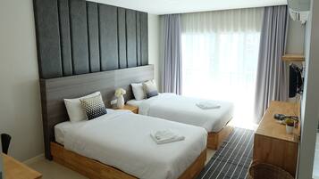 A hotel room with two beds, a wooden desk, a small table, a wall-mounted TV, and a window with curtains.