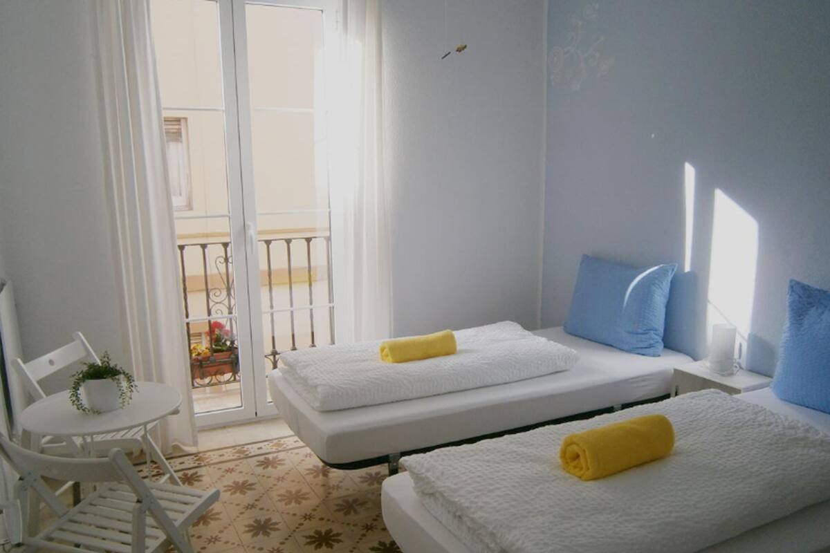 A hotel room with two beds, a small table with a chair, and a balcony with a railing.