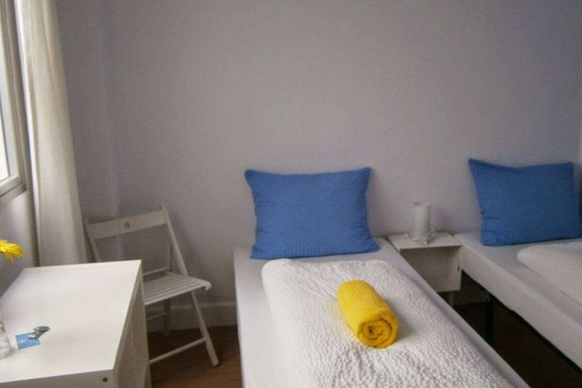 A small, simple room with two single beds, each with a blue pillow and a yellow towel. There is a small white desk with a chair and a window to the left.