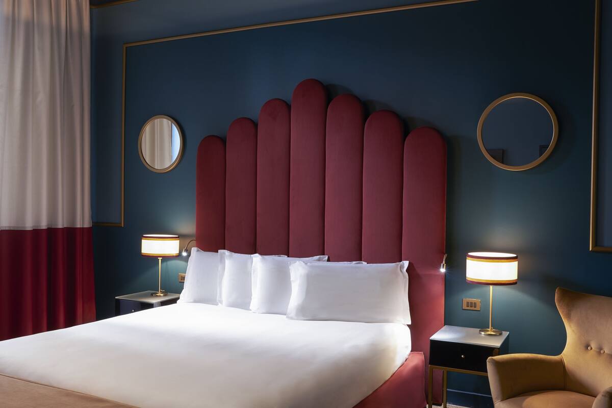 A hotel room with a large bed, two bedside tables with lamps, a red upholstered headboard, and a round mirror on the wall.