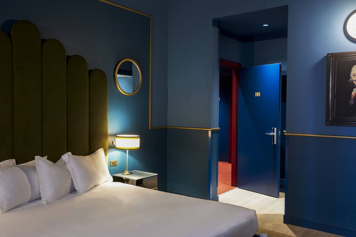 A hotel room with a bed, bedside table, lamp, and a door leading to another room.