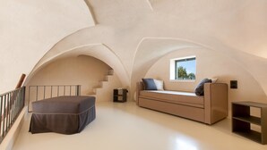 Une chambre spacieuse située dans les combles, avec un plafond haut, un canapé intégré, une petite table et une fenêtre donnant sur le ciel.