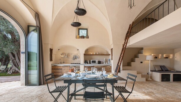 Une salle à manger spacieuse avec un haut plafond en arc, une table centrale dressée pour le repas, et une vue sur la cuisine et l’espace repas extérieur.