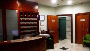 Zona de recepción de hotel con un mostrador de madera, un tablón de anuncios con etiquetas de habitaciones, una silla verde y un letrero que indica la salida.