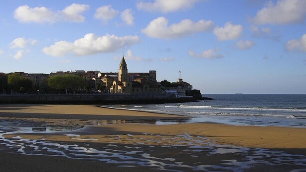 Paisaje costero con playa arenosa, un edificio grande con torre y el océano al fondo.