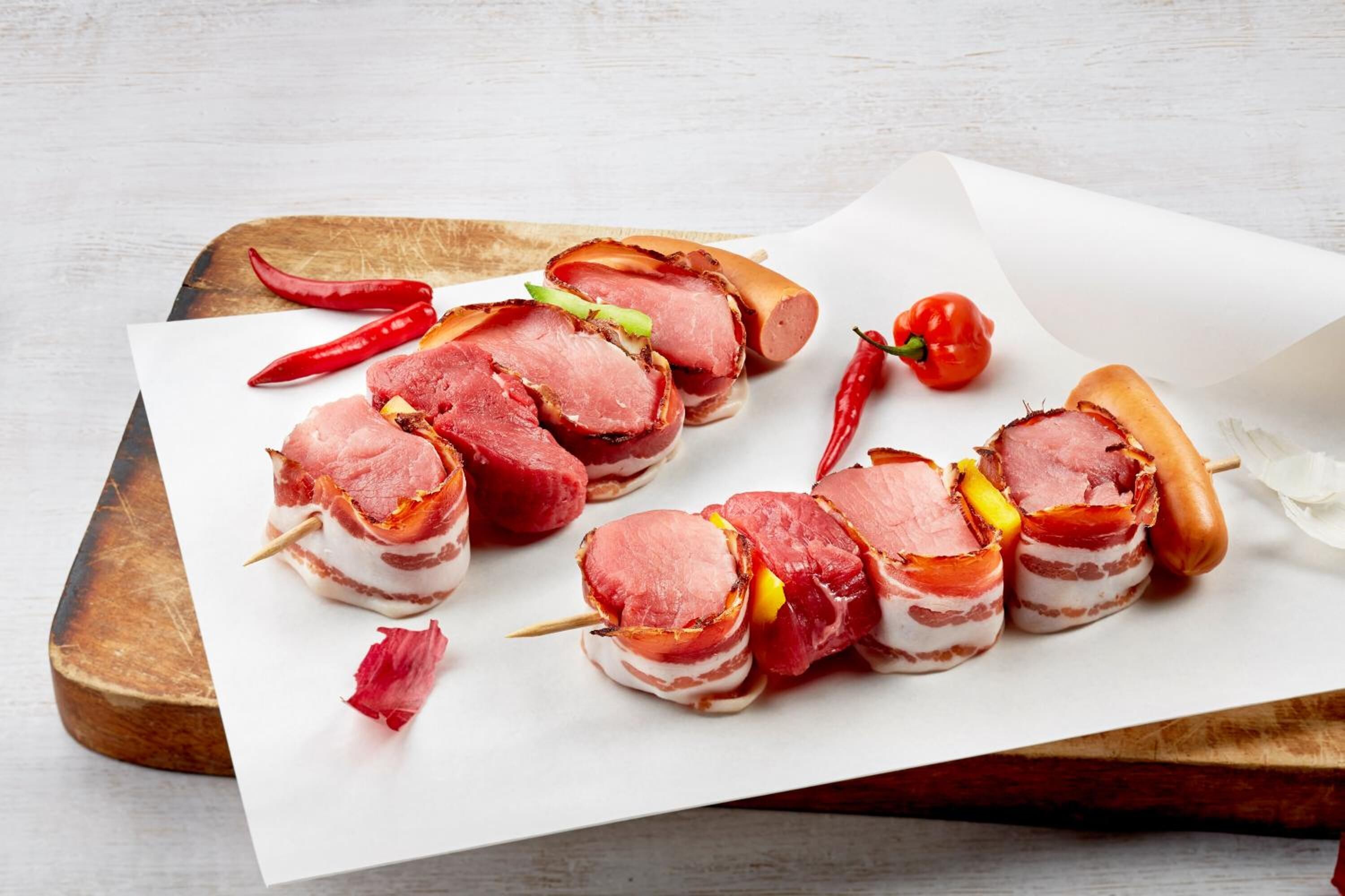 Fleischspieße in Speck gehüllt auf einer Holzplanke.