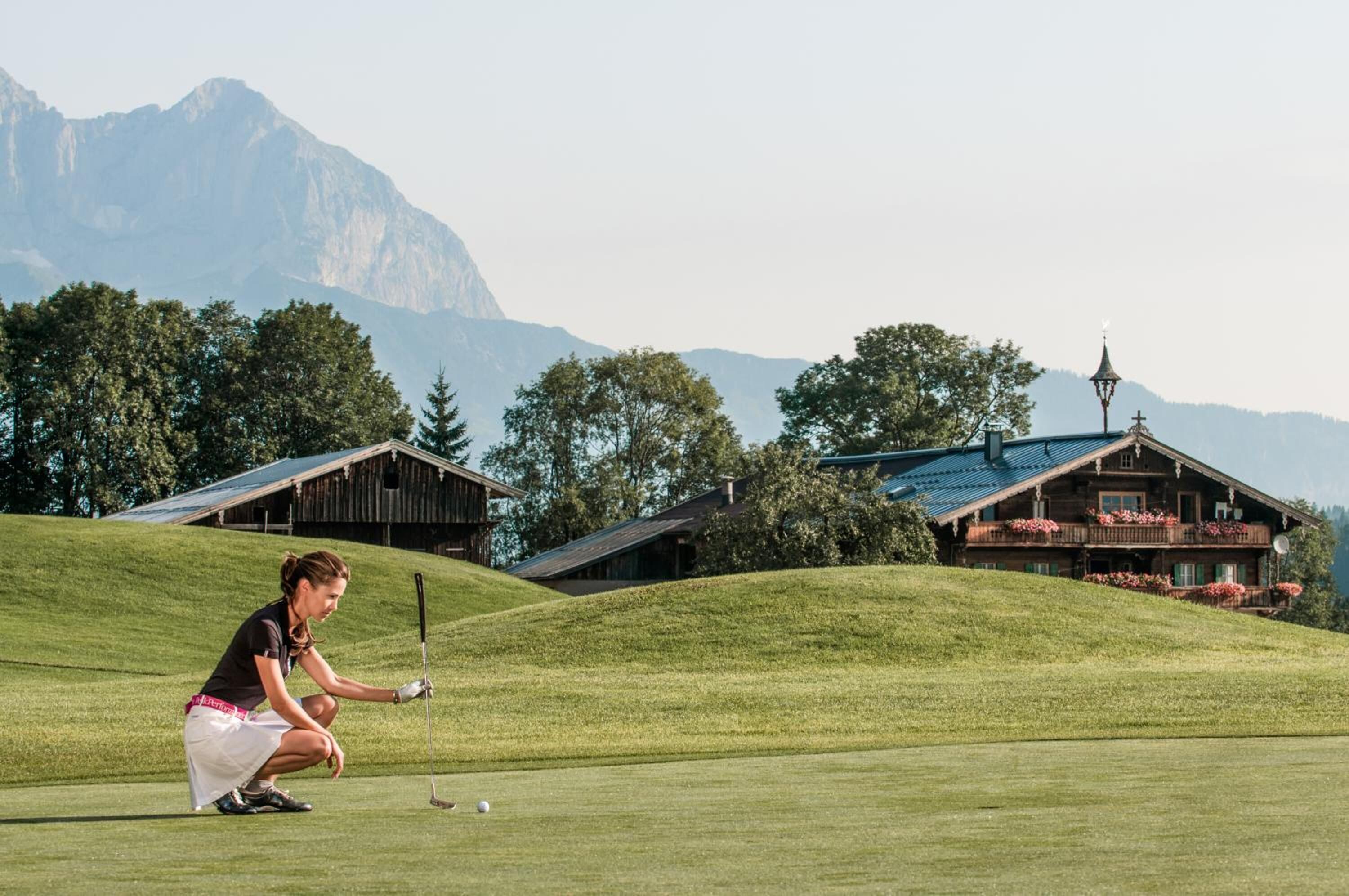 Eine Frau spielt Golf auf einem gepflegten Golfplatz mit einer Bergkulisse und traditionellen Holzhäusern.