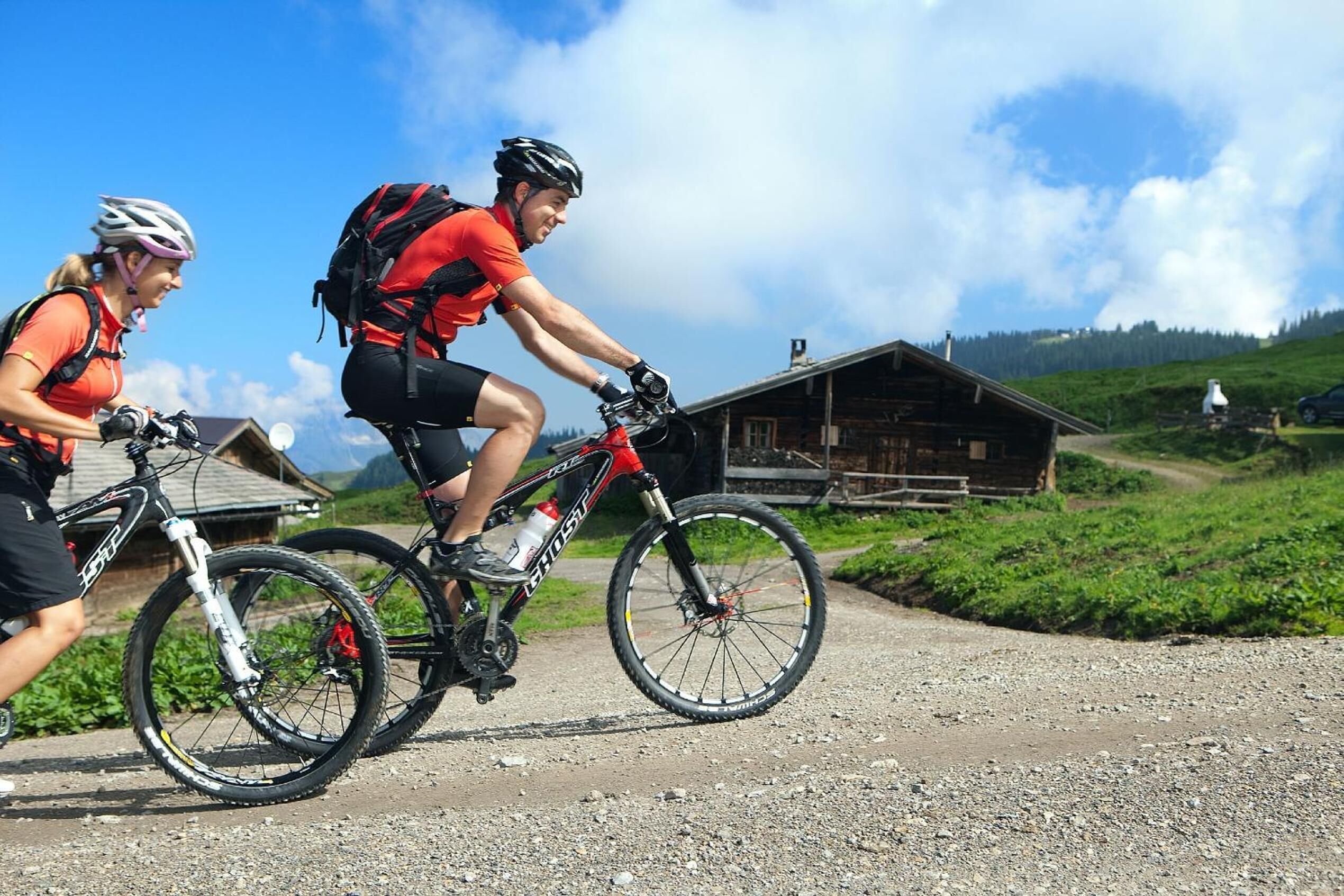 Deux cyclistes sur un chemin de gravier, avec une cabane en bois en arrière-plan.