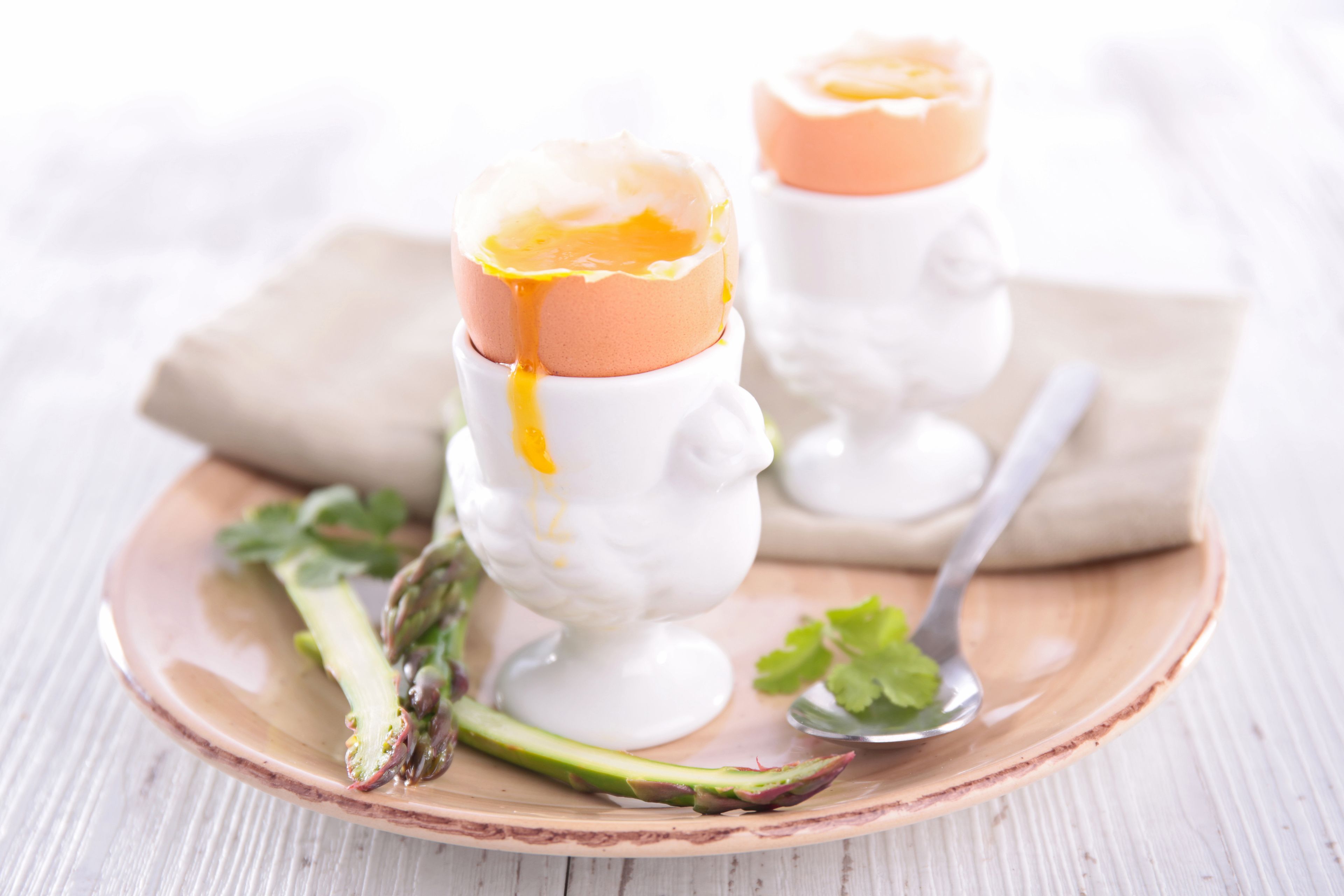 Deux coquetiers contenant chacun un œuf dur, disposés sur une assiette avec des asperges et une cuillère.