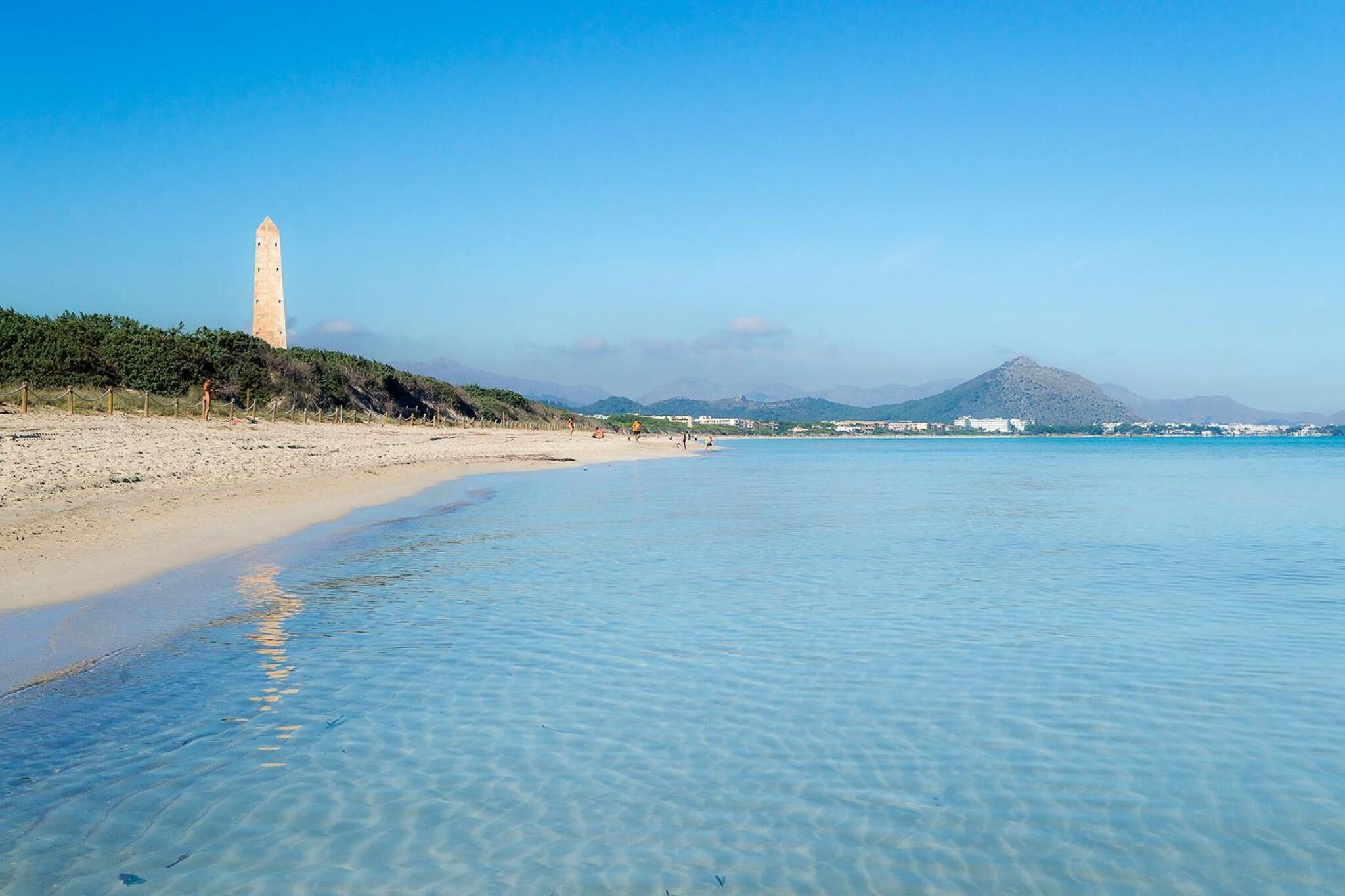 Ein Strand mit klarem Wasser, einem Leuchtturm und Bergen im Hintergrund.
