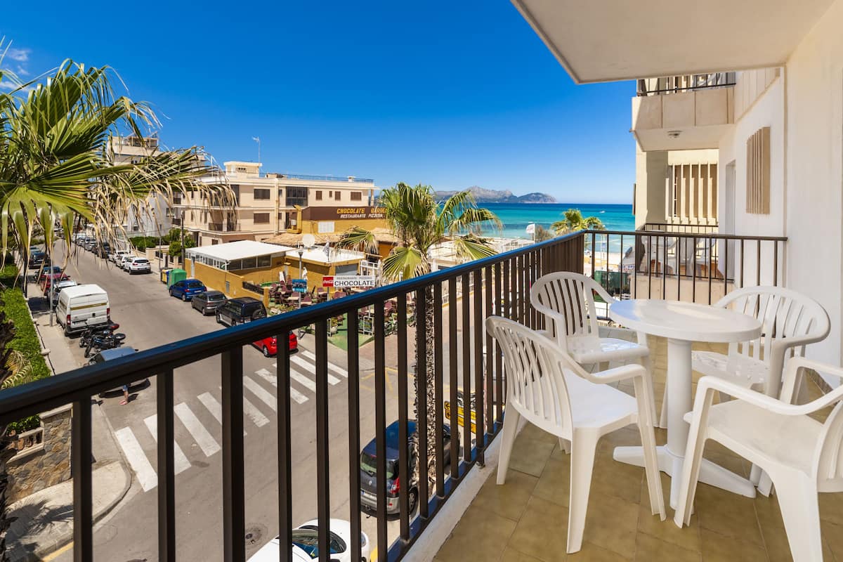 Un balcon avec des chaises et une table en plastique blanc, donnant sur une rue où sont garées des voitures et sur une plage, avec des bâtiments en arrière-plan.