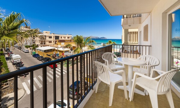 Un balcón con sillas y una mesa de plástico blanco, con vista a una calle con coches aparcados y una playa con edificios al fondo.