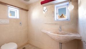 Ein Badezimmer mit einer beige gefärbten Wand, einer weißen Toilette, einem beigen Waschbecken und einem Fenster mit Blick auf ein Gebäude.