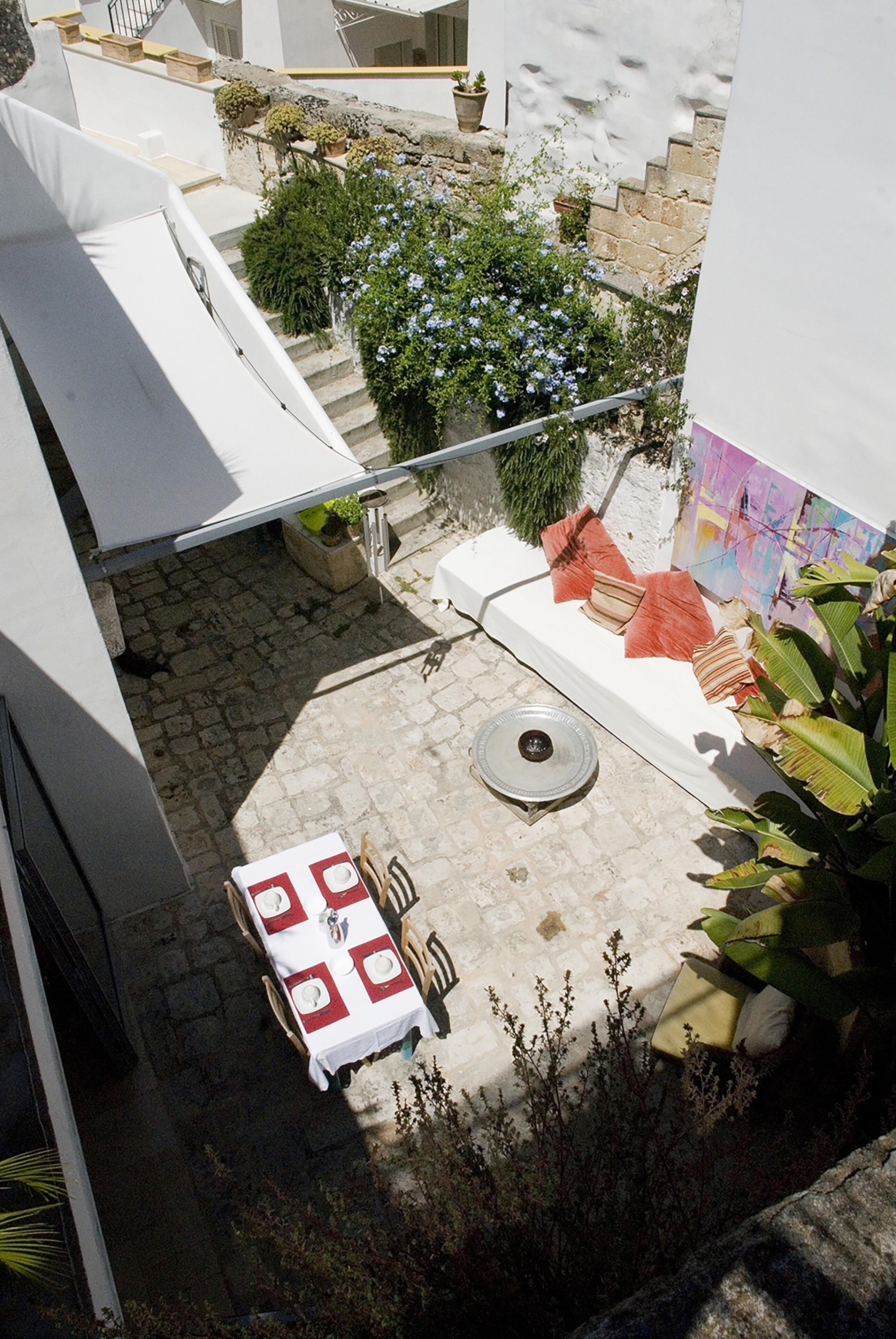 Un patio con una mesa de comedor preparada para cuatro personas, rodeado de plantas en macetas y una escalera que conduce a un nivel superior.