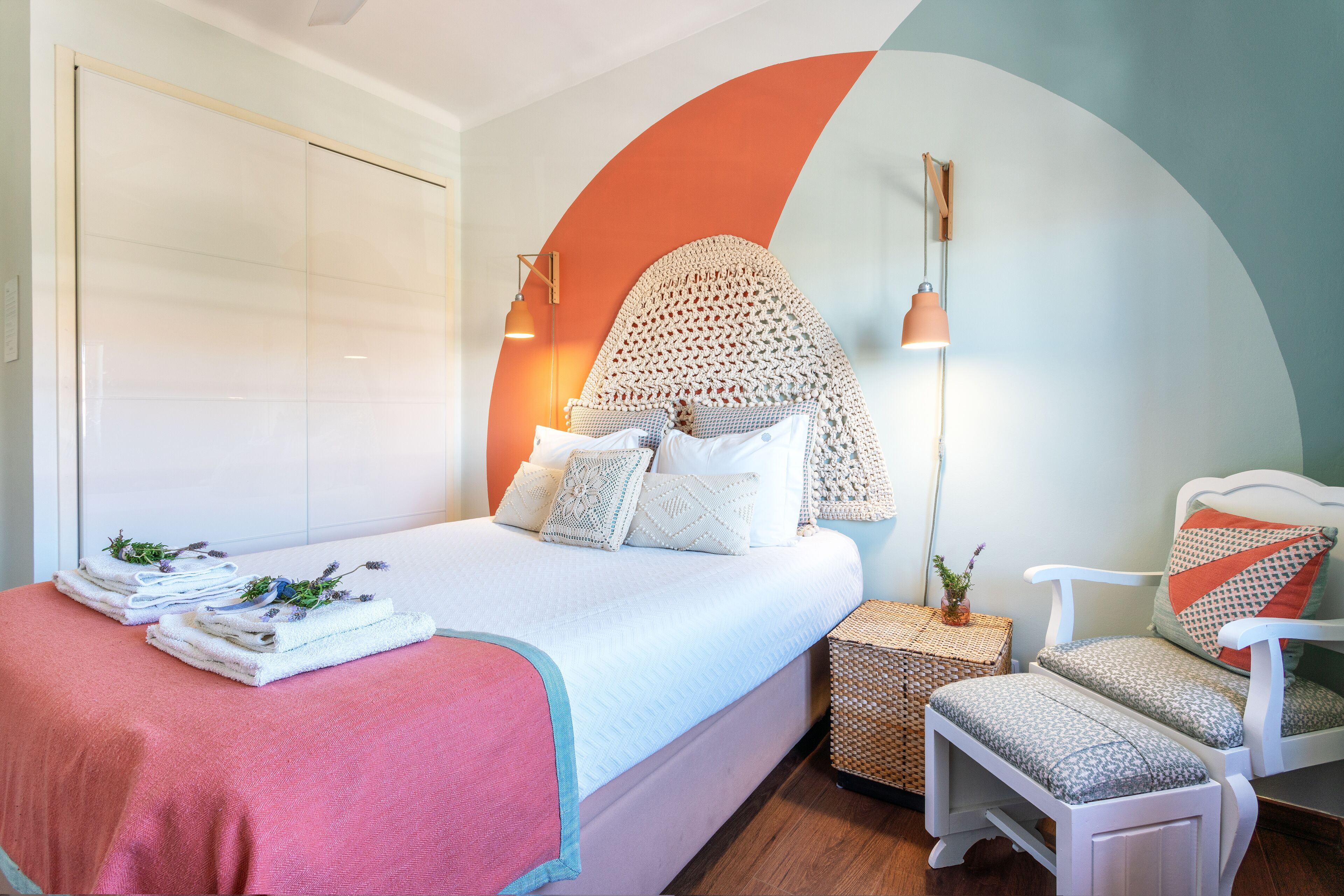 Un lit bien fait, recouvert d’une couverture rose, une chaise blanche avec des coussins à motifs et une table de chevet sur laquelle repose un panier en osier.