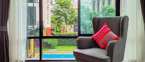 A modern room with a large window, a grey armchair with a red and black striped pillow, and a view of a building and greenery outside.
