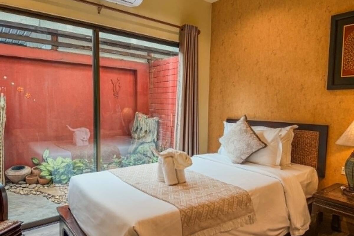 Hotelkamer met een bed, nachtkastje, lamp, raam en uitzicht op een rode muur en planten buiten.