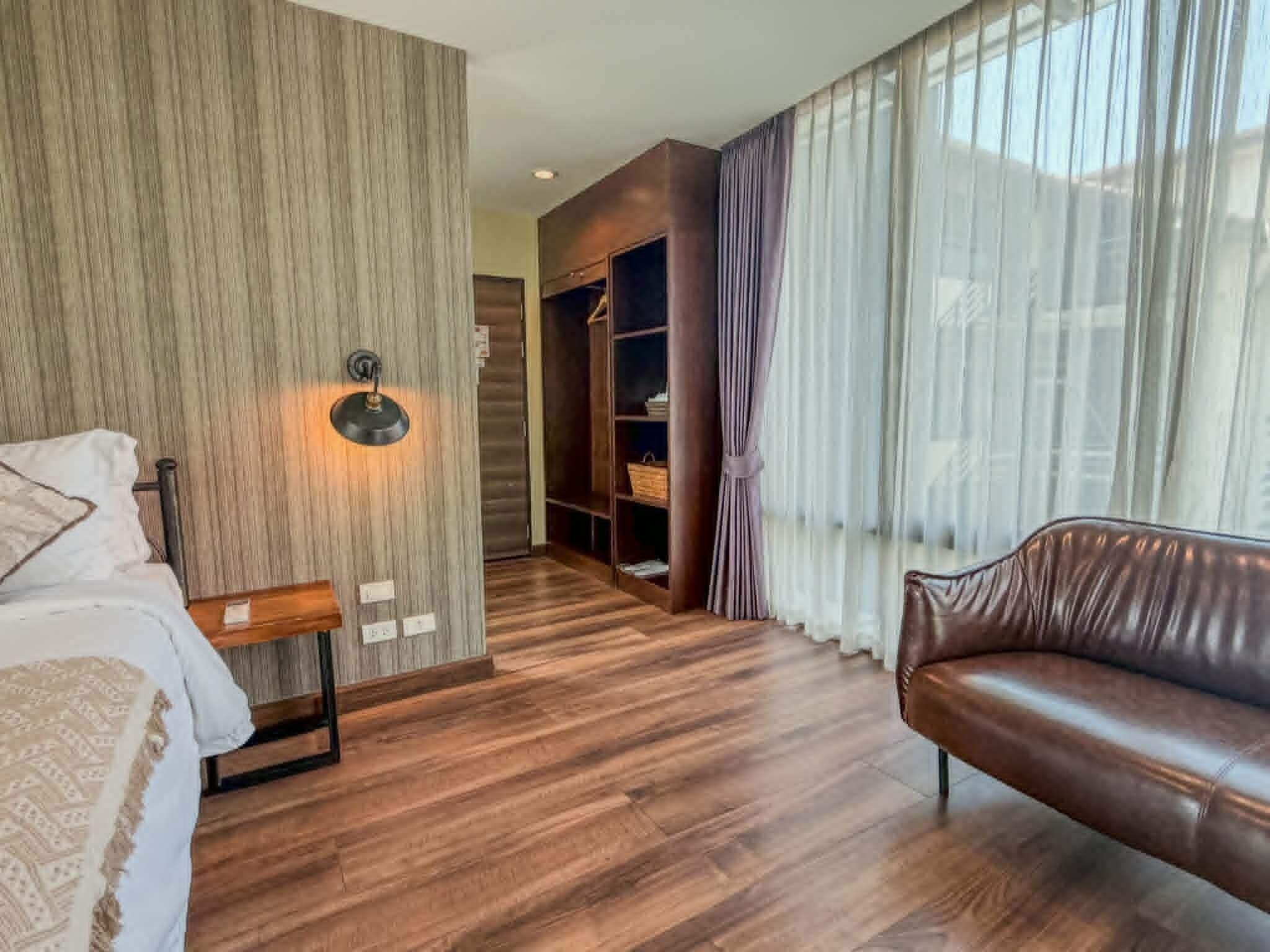 A hotel room with a bed, a wooden floor, a leather sofa, a wardrobe, and a large window with curtains.