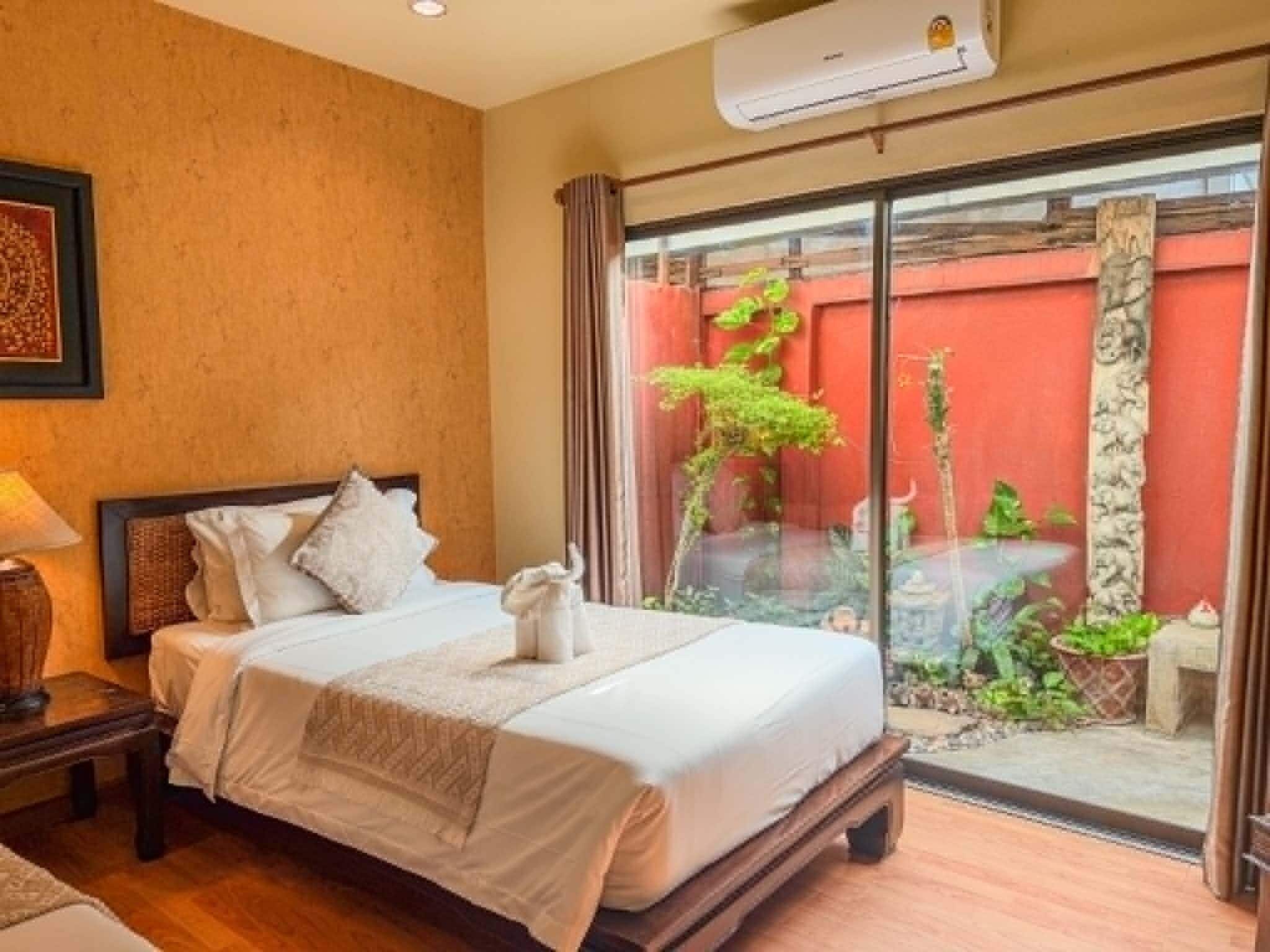 A hotel room with a bed, a nightstand, a lamp, and a view of a red wall and greenery outside.