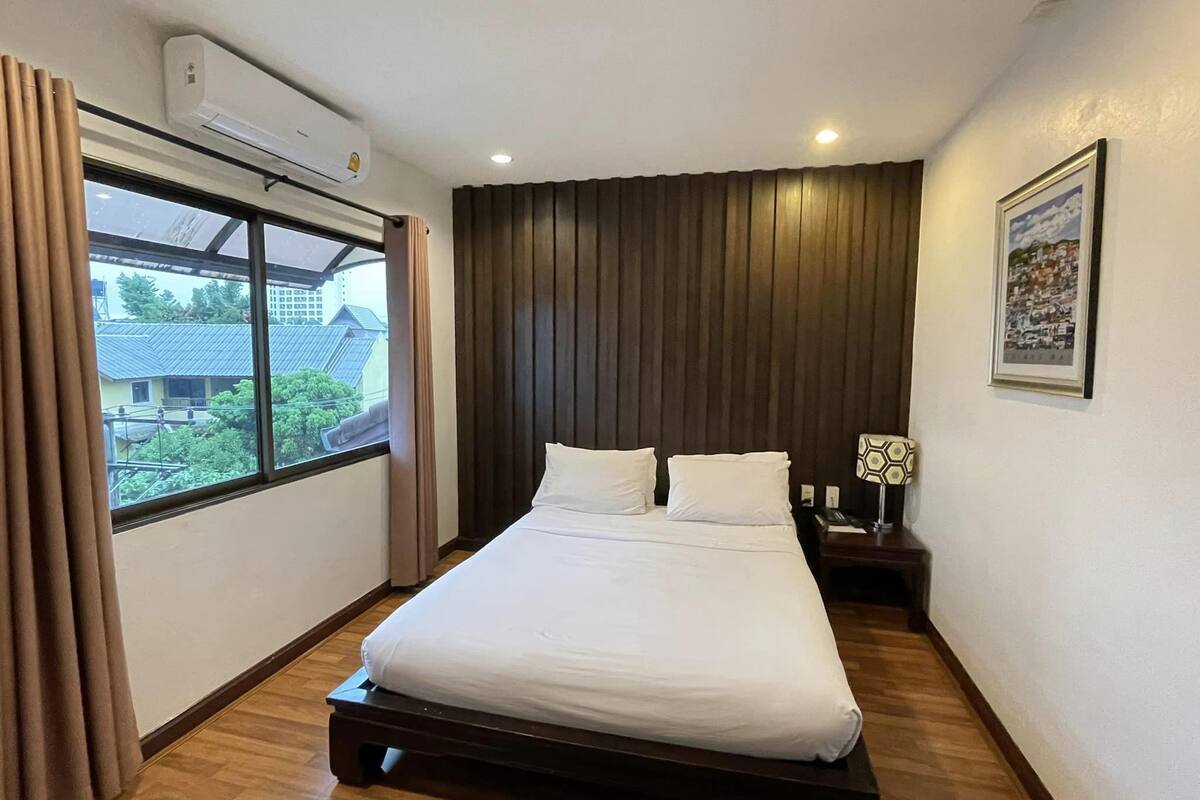 Hotelkamer met een groot bed, een raam met gordijnen, een airconditioningapparaat, een nachtkastje met een lamp en een ingelijst foto aan de muur.