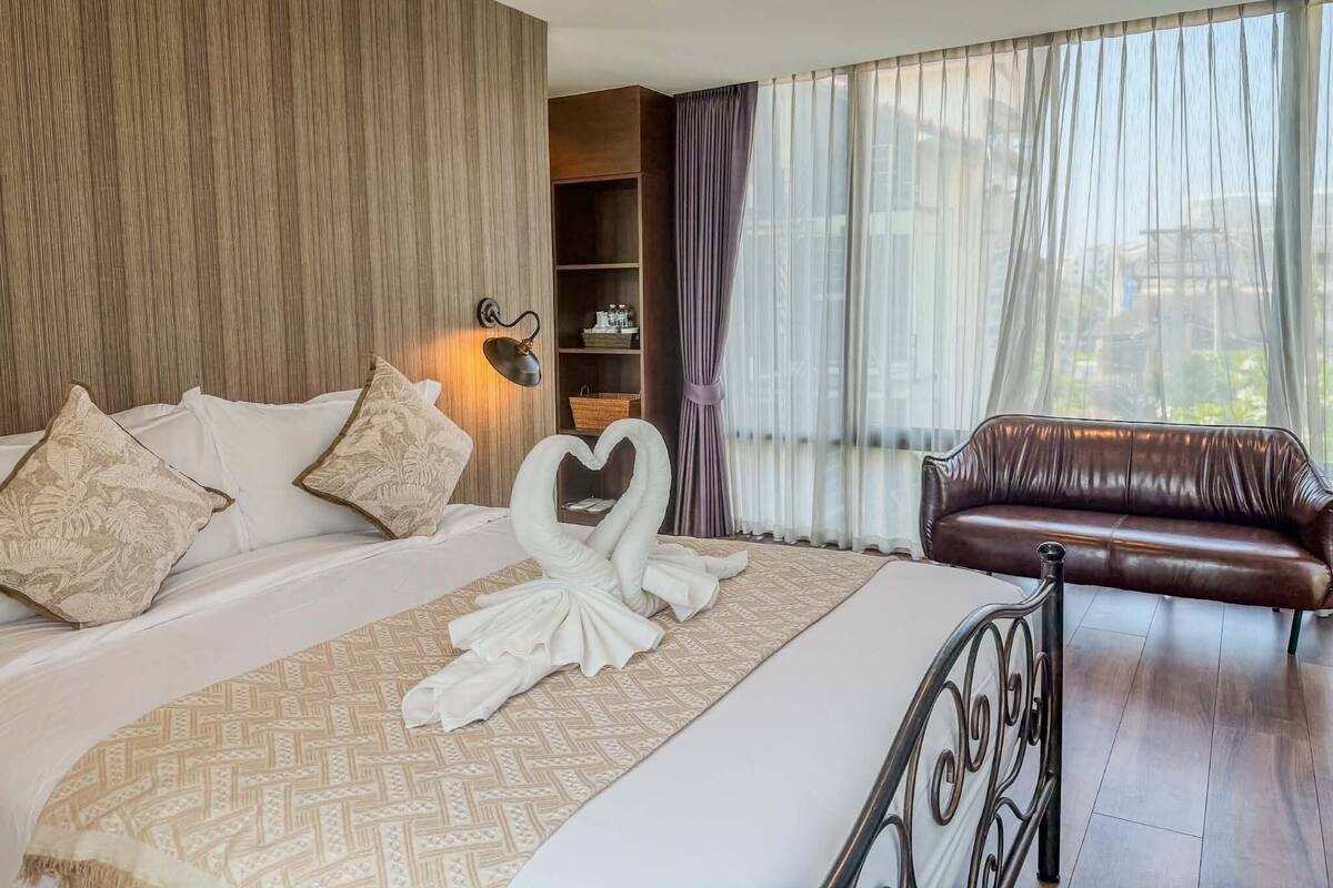 Een hotelkamer met een bed, een handdoek in de vorm van een hart, een bruine leren bank, een wandlamp en een groot raam met dunne gordijnen.