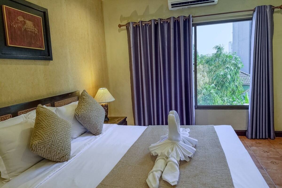 Hotelkamer met een bed, kussens, een nachtkastje met een lamp, een raam met gordijnen en een airconditioning.