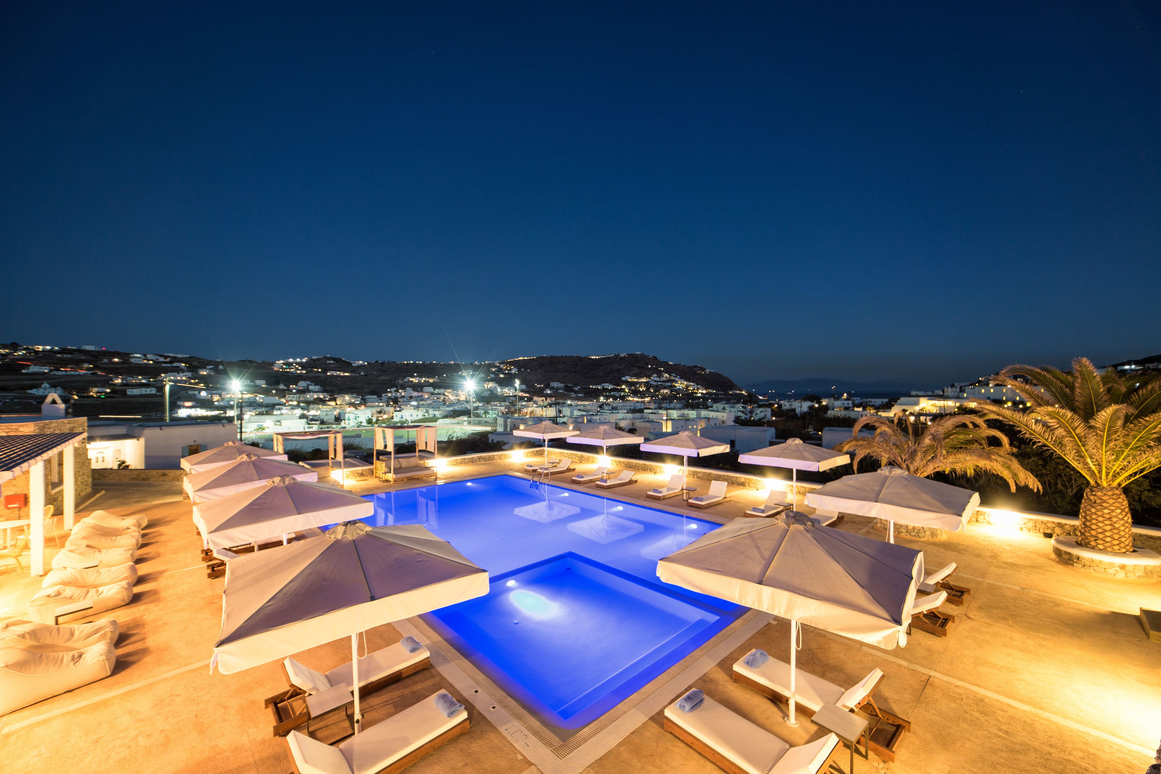 A nighttime view of a rooftop pool with lounge chairs and umbrellas, overlooking a cityscape.