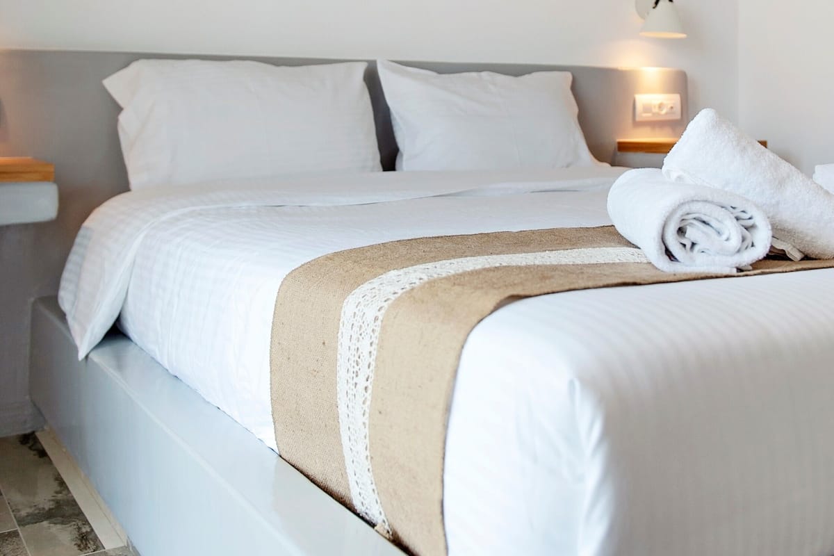 A neatly made bed with a white and beige bedspread, two white pillows, and a rolled towel on the side.