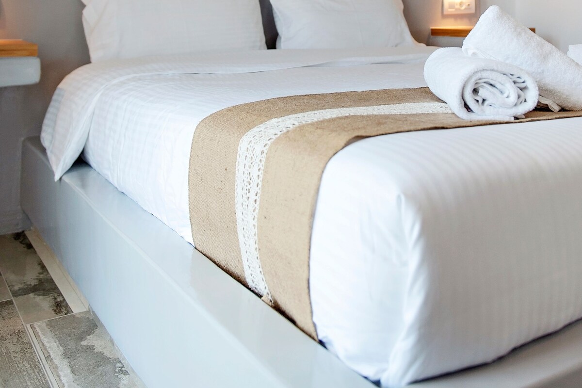 A neatly made bed with a white and beige bedspread, two white pillows, and a rolled towel on the side.