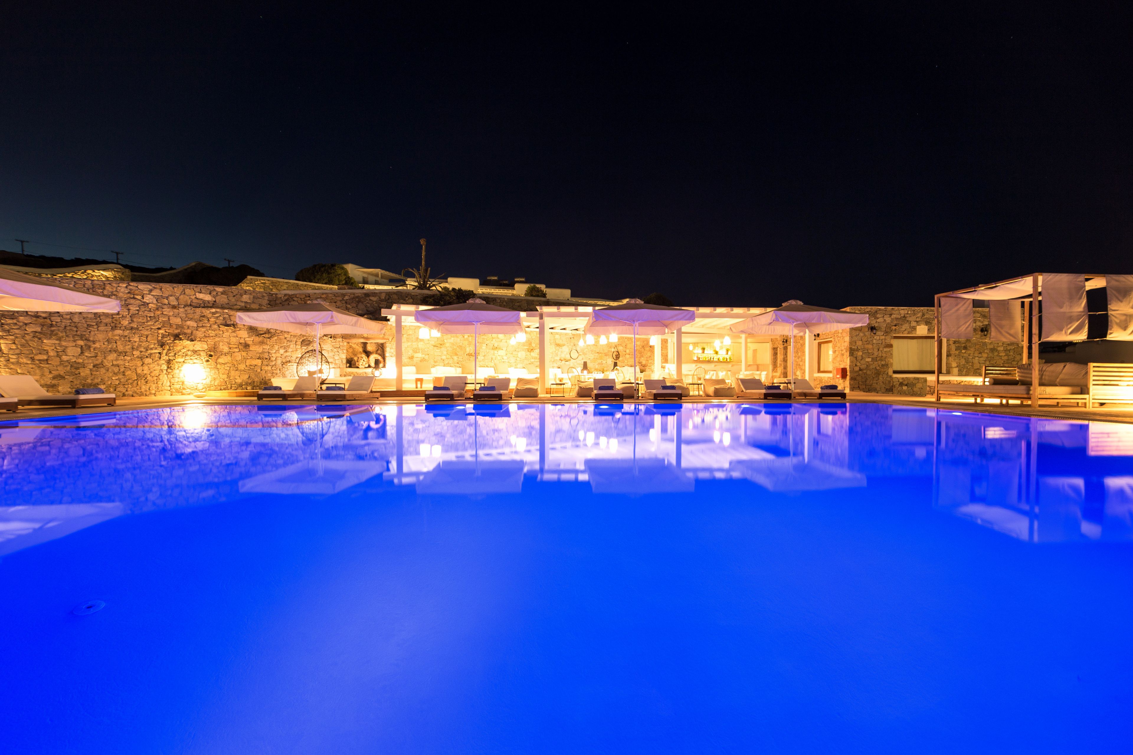 A nighttime swimming pool with lounge chairs and umbrellas, surrounded by stone walls and a building with illuminated windows.