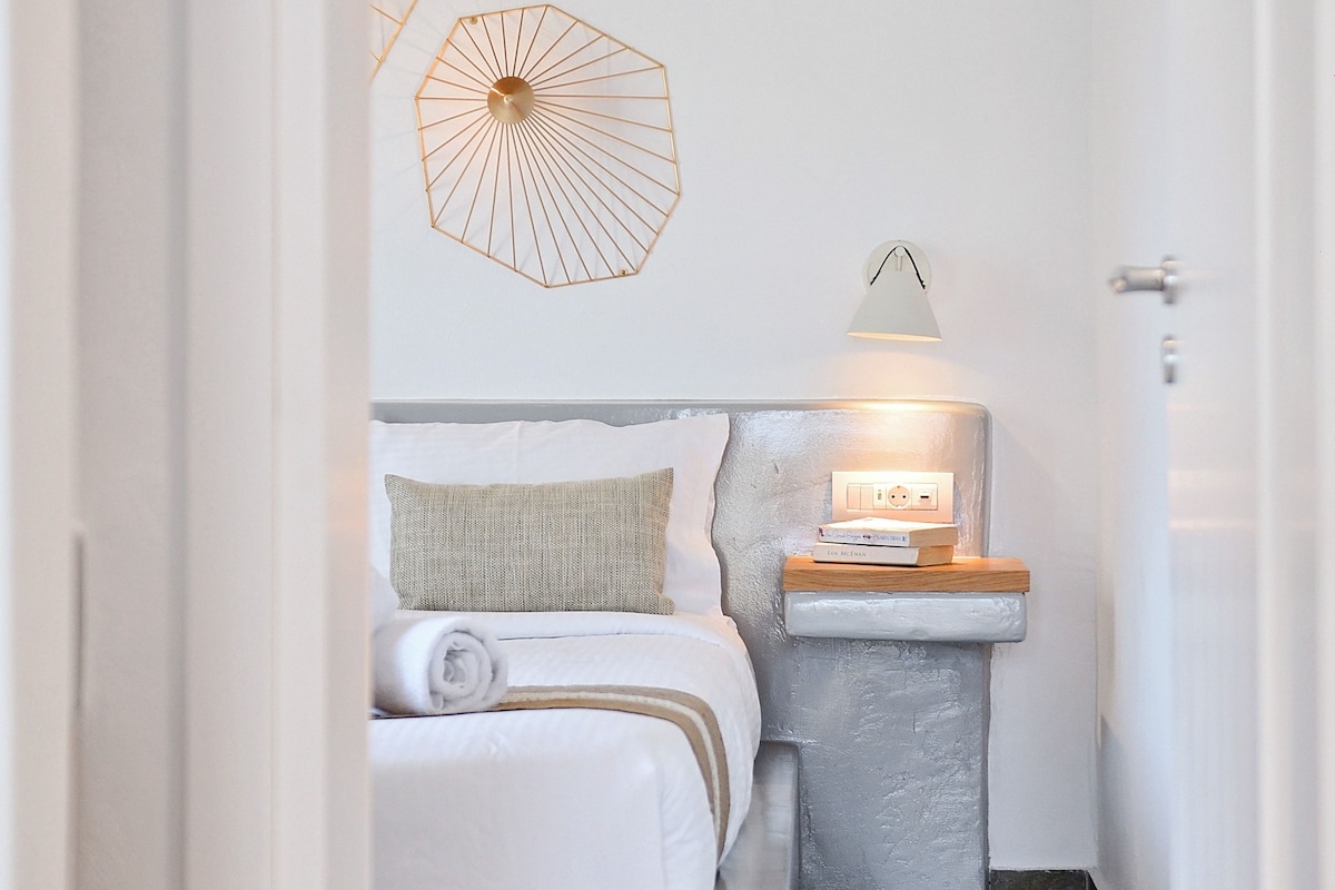 Um corredor estreito que dá para um quarto com uma cama, mesa de cabeceira, luminária na parede e um guarda-chuva decorativo.