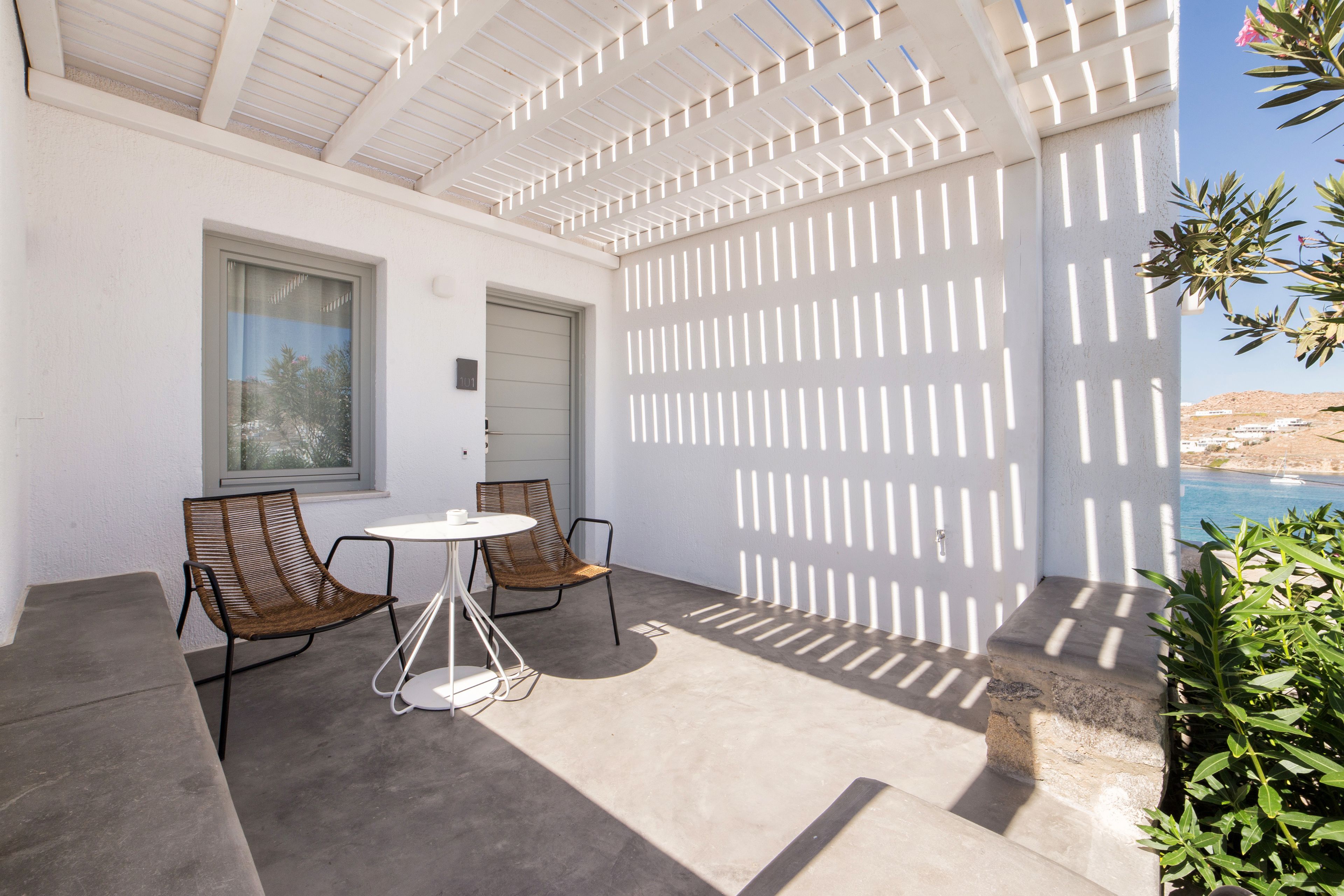 Un patio con una pérgola blanca, dos sillas de mimbre, una mesita redonda y vistas a una piscina y jardines.