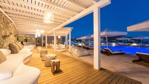A wooden deck with lounge chairs and a pool area, featuring a stone wall and white furniture. - Osom Resort (Mykonos)