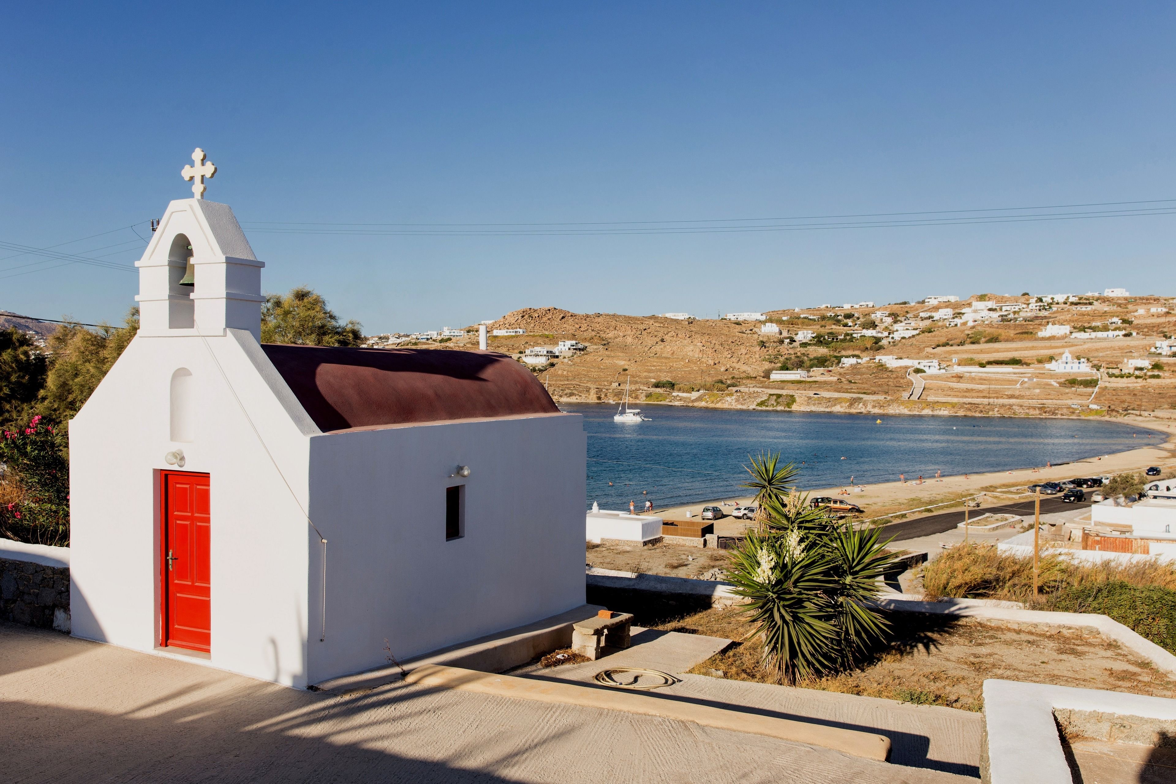 A white church with a red door and a bell tower, overlooking a coastal village with houses and a beach.