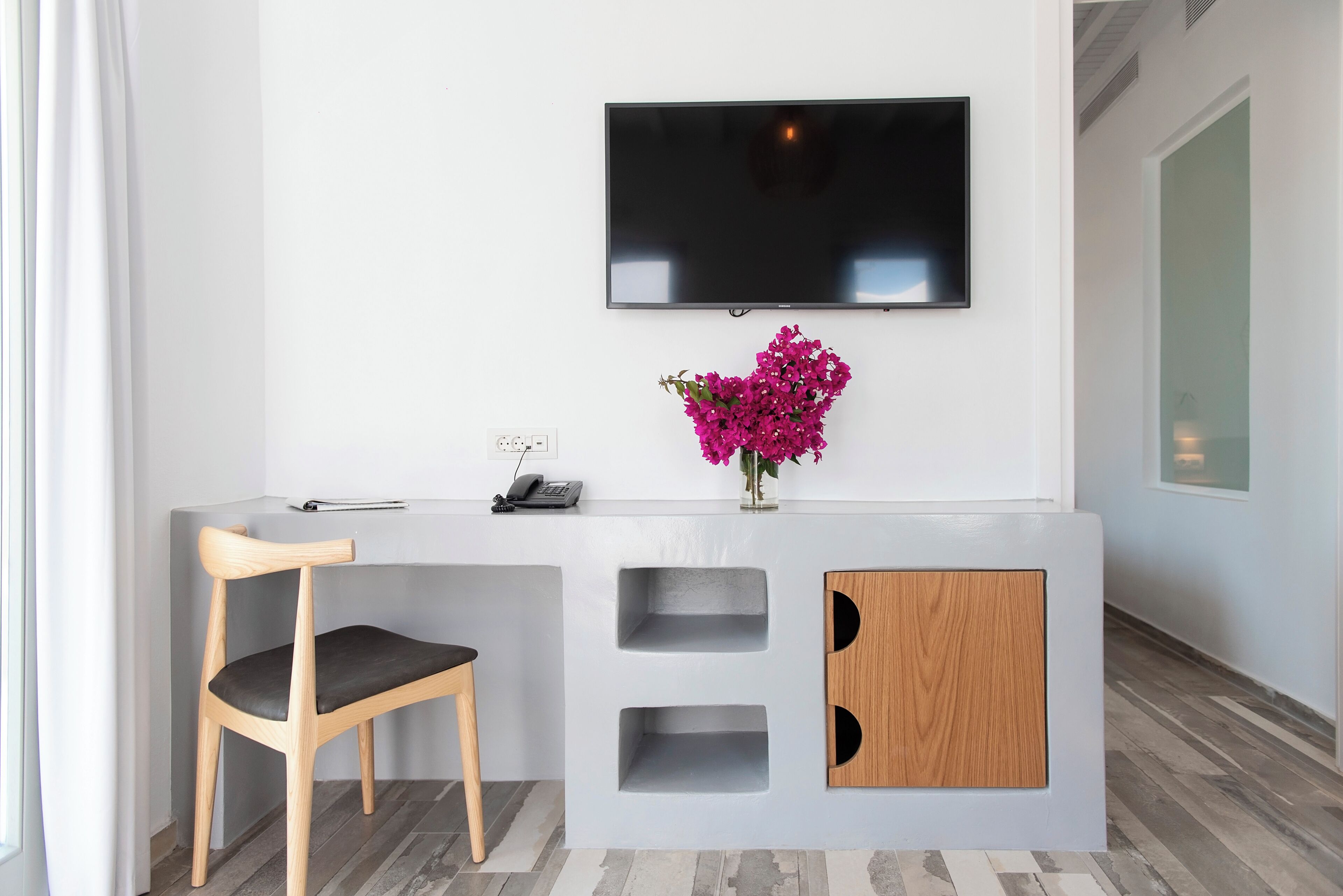 Une chambre d’hôtel moderne, dotée d’une chaise en bois, d’un téléviseur à écran plat fixé au mur et d’une petite armoire en bois avec des étagères.