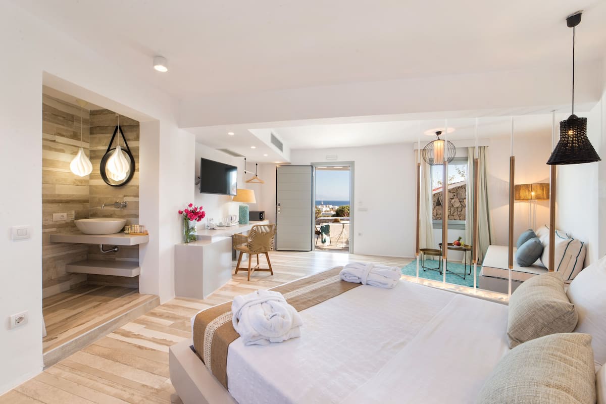 A modern hotel room with a large bed, a bathroom with a sink and mirror, a dining area with a table and chairs, and a view of the sea.