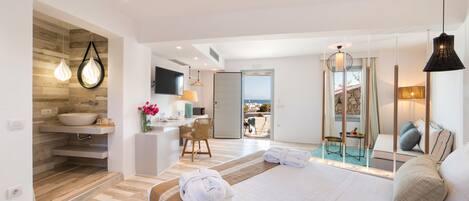 A modern hotel room with a large bed, a bathroom with a sink and mirror, a dining area with a table and chairs, and a view of the sea.